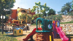 A playground with a slide and climbing structures, surrounded by greenery and a building in the background.