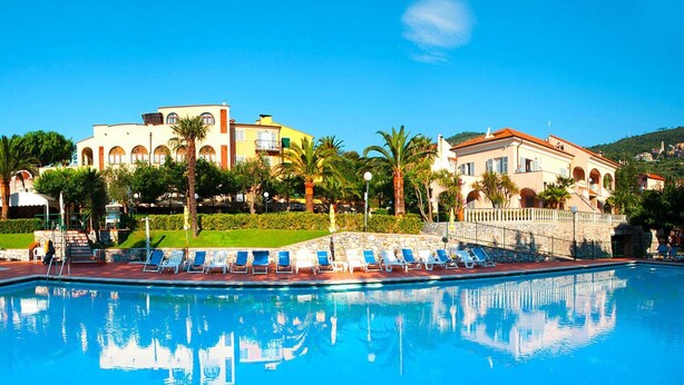A swimming pool with lounge chairs, palm trees, and a building with arched windows in the background.