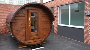 A wooden barrel-shaped structure with a door, situated on a paved area with a brick building in the background. - Ferienwohnung in Westerland (Sylt)