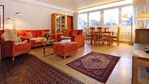 A living room with a red sofa set, a coffee table, a patterned rug, and a dining area with wooden chairs and a table. - Ferienwohnung in Westerland (Sylt)