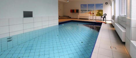 Sisätiloissa sijaitseva uima-allas, jonka seinät ja lattia ovat laatoitettu, siinä on penkki ja ikkunassa sälekaihtimet.