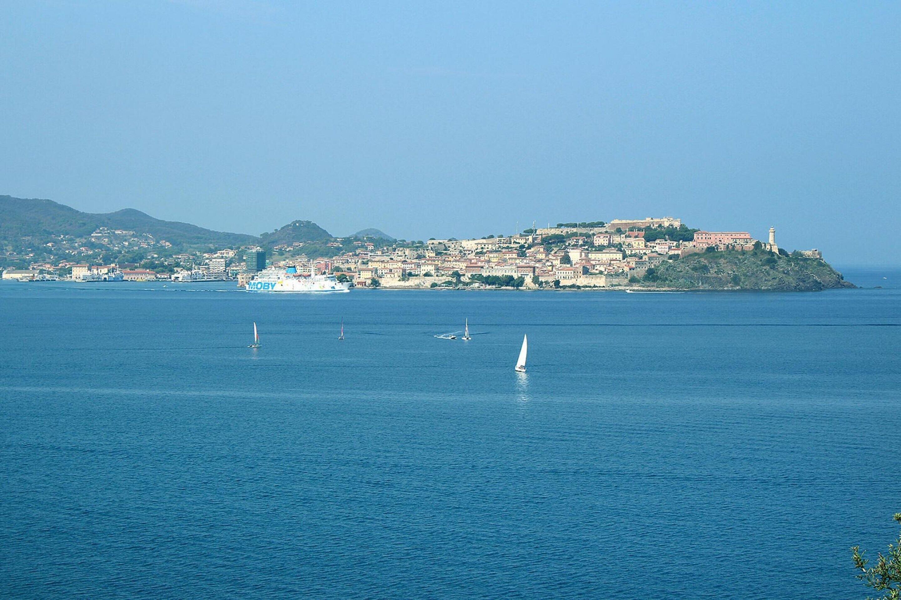 A coastal town with a prominent castle on a hill, calm sea, and sailboats.
