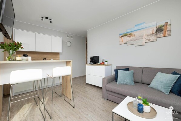 A modern living room with a grey sofa, white chairs, and a coffee table. A kitchen area with white cabinets and a wooden countertop is visible. - Apartment Very Close to the Seaside Beach (Rewal)