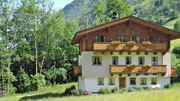 Una cabaña de madera de dos pisos con balcones, rodeada de abundante vegetación y montañas.