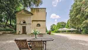 A rustic outdoor dining area with a table and chairs, a small building with a door, and a gravel ground. - Country House in Tuscany With Pool and Olive Grove (Capannoli)