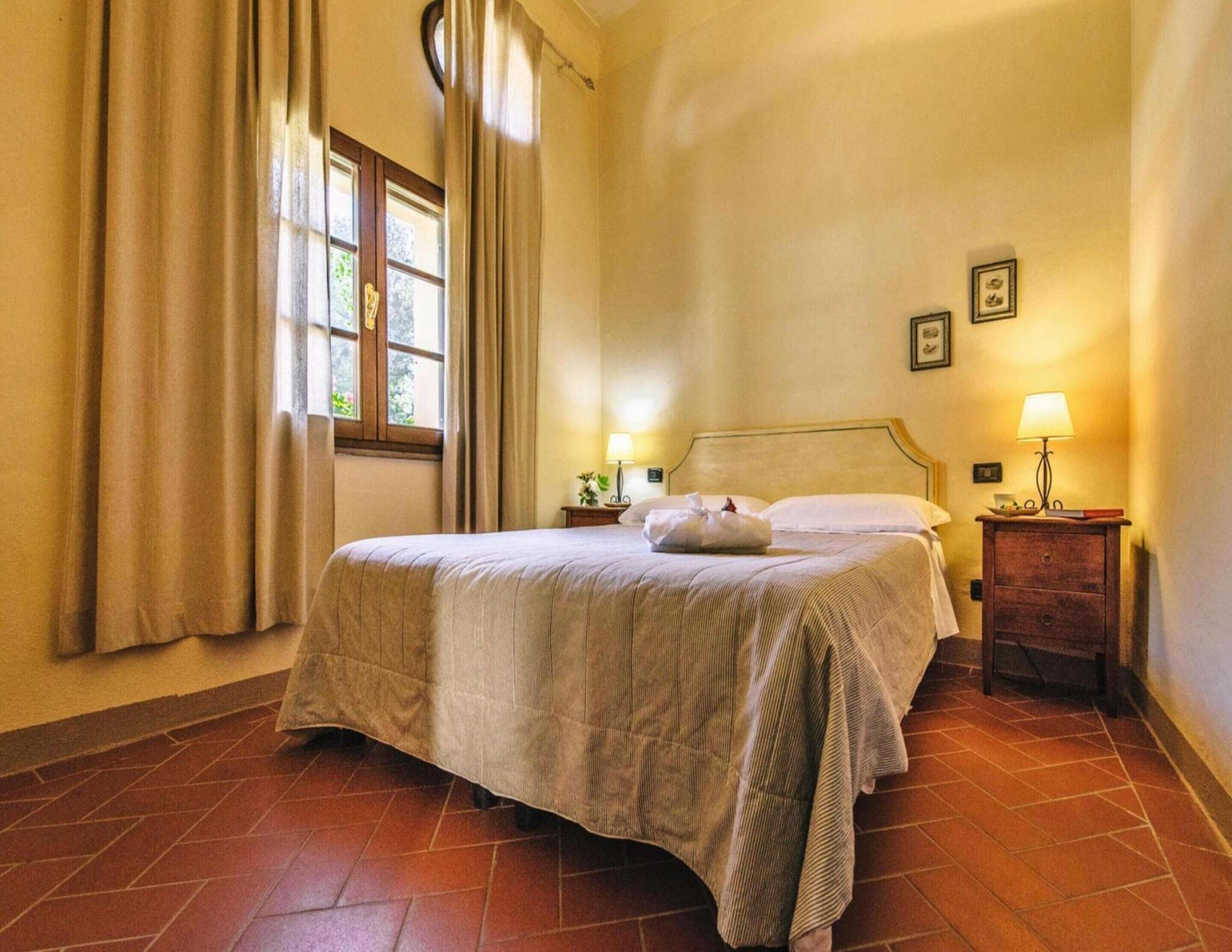 A bedroom with a bed, curtains, a window, a bedside table with a lamp, and a wall-mounted picture.