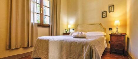 A bedroom with a bed, curtains, a window, a bedside table with a lamp, and a wall-mounted picture.