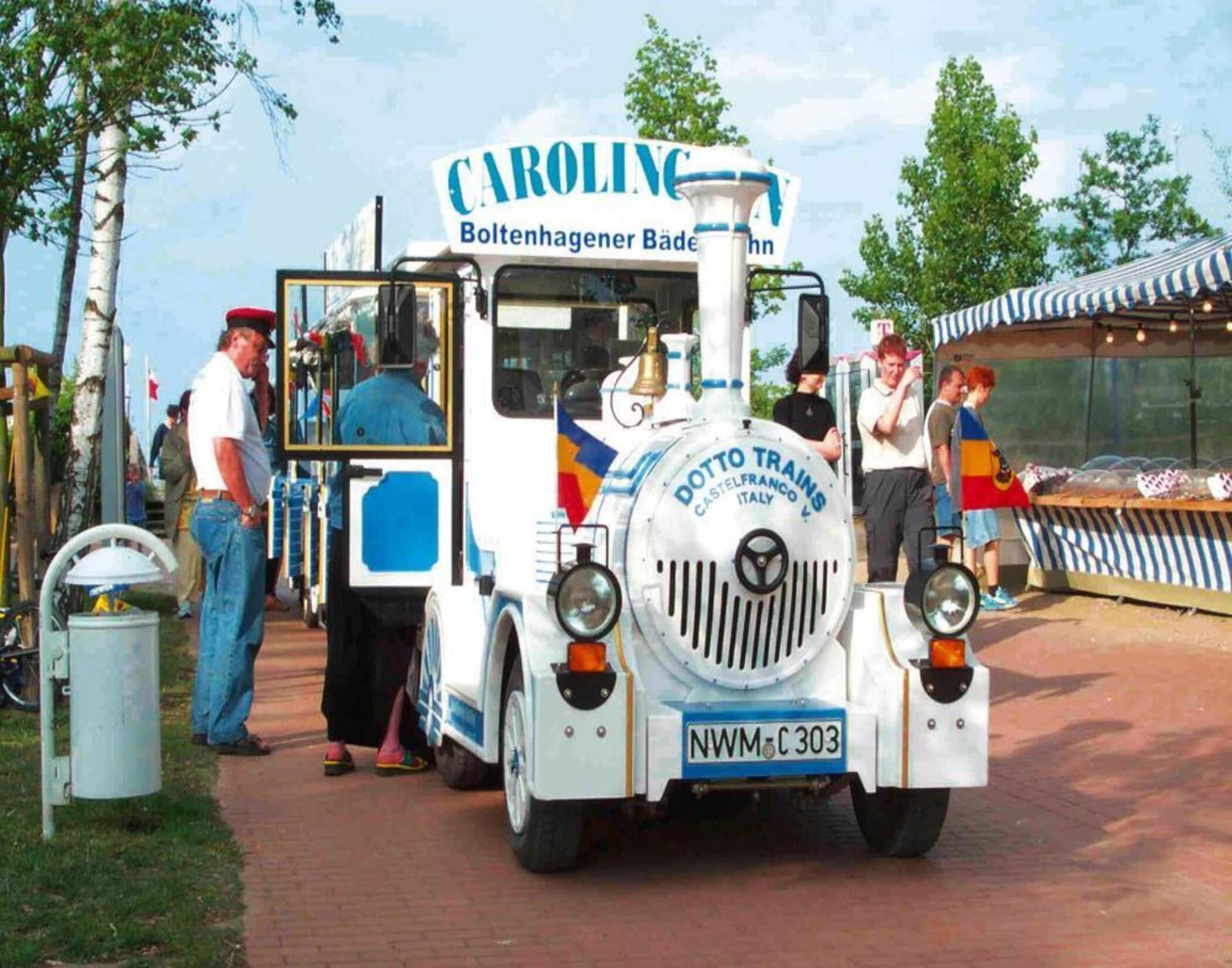 A vintage train with "Caroline" and "Dotto Trains" branding, parked at a market with people around.