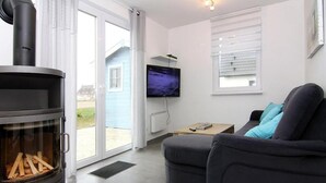 Ein modernes Wohnzimmer mit Kaminofen, einem grauen Sofa, einem Fernseher und Blick auf ein Haus durch die Glastür.
