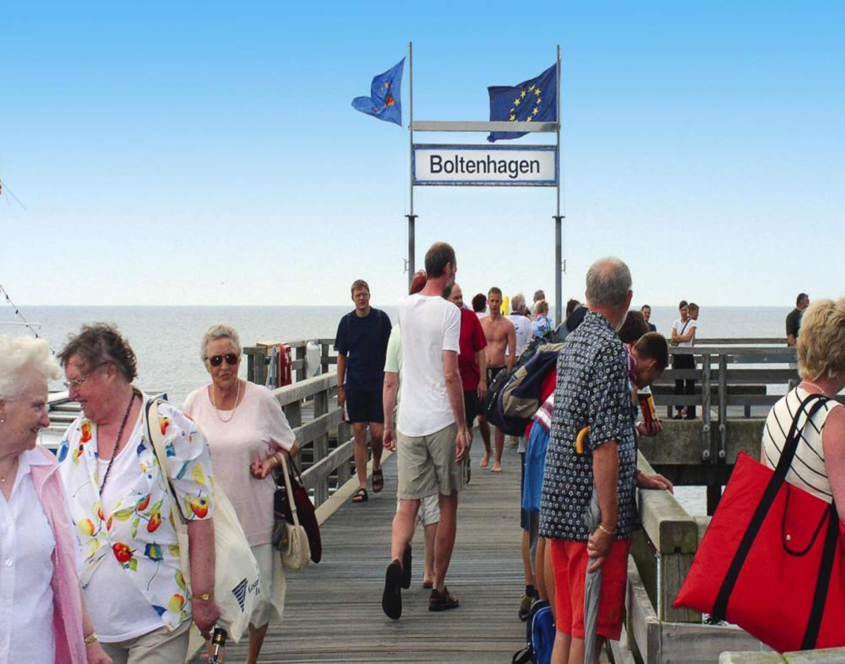 Un muelle con gente caminando, un cartel que dice "Boltenhagen", y banderas de la Unión Europea y de Alemania.