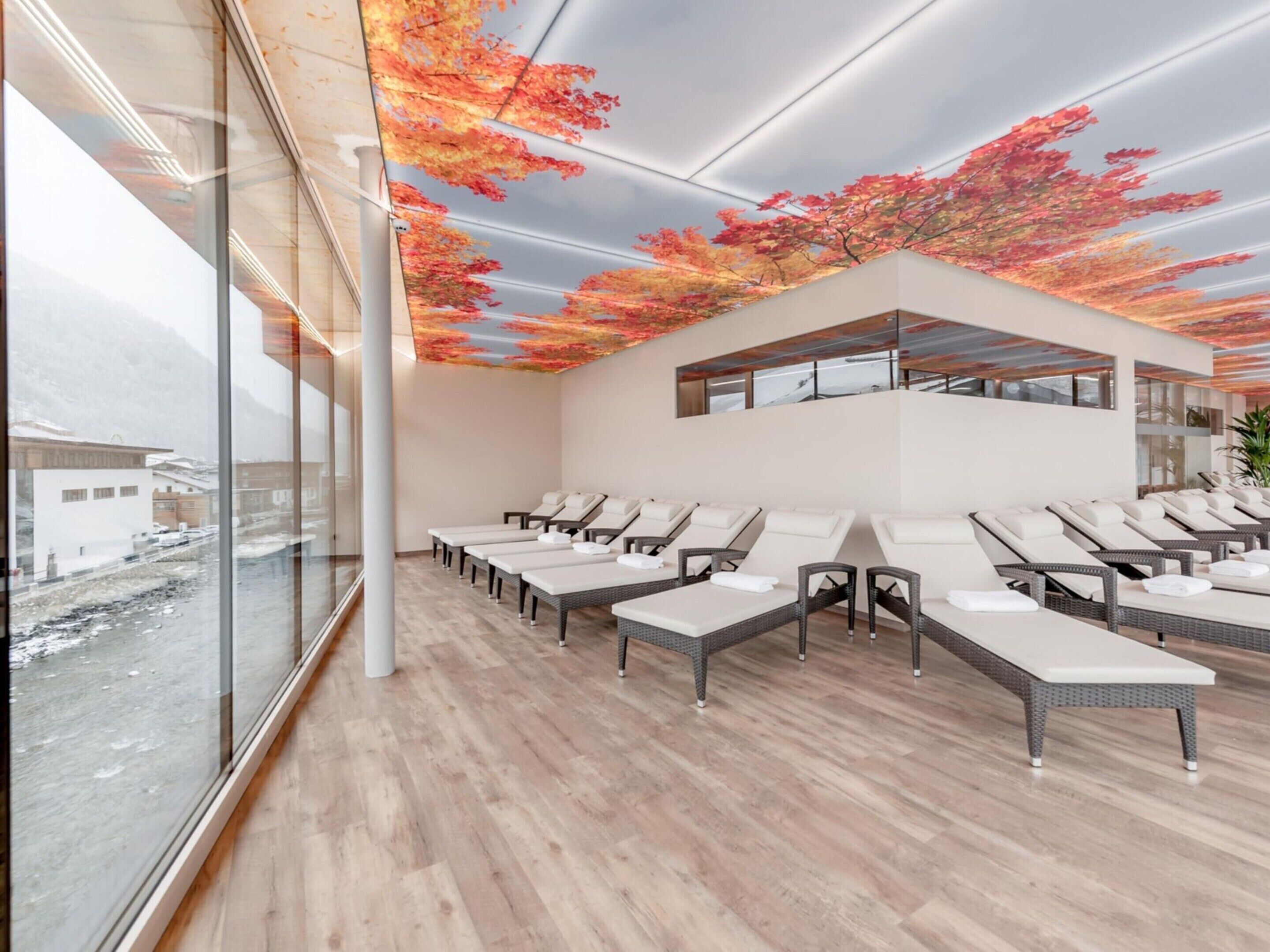 Ein moderner Wellnessraum mit Wandgemälde von Herbstlaub, einer Reihe von Liegestühlen und Blick auf eine verschneite Landschaft durch große Fenster.