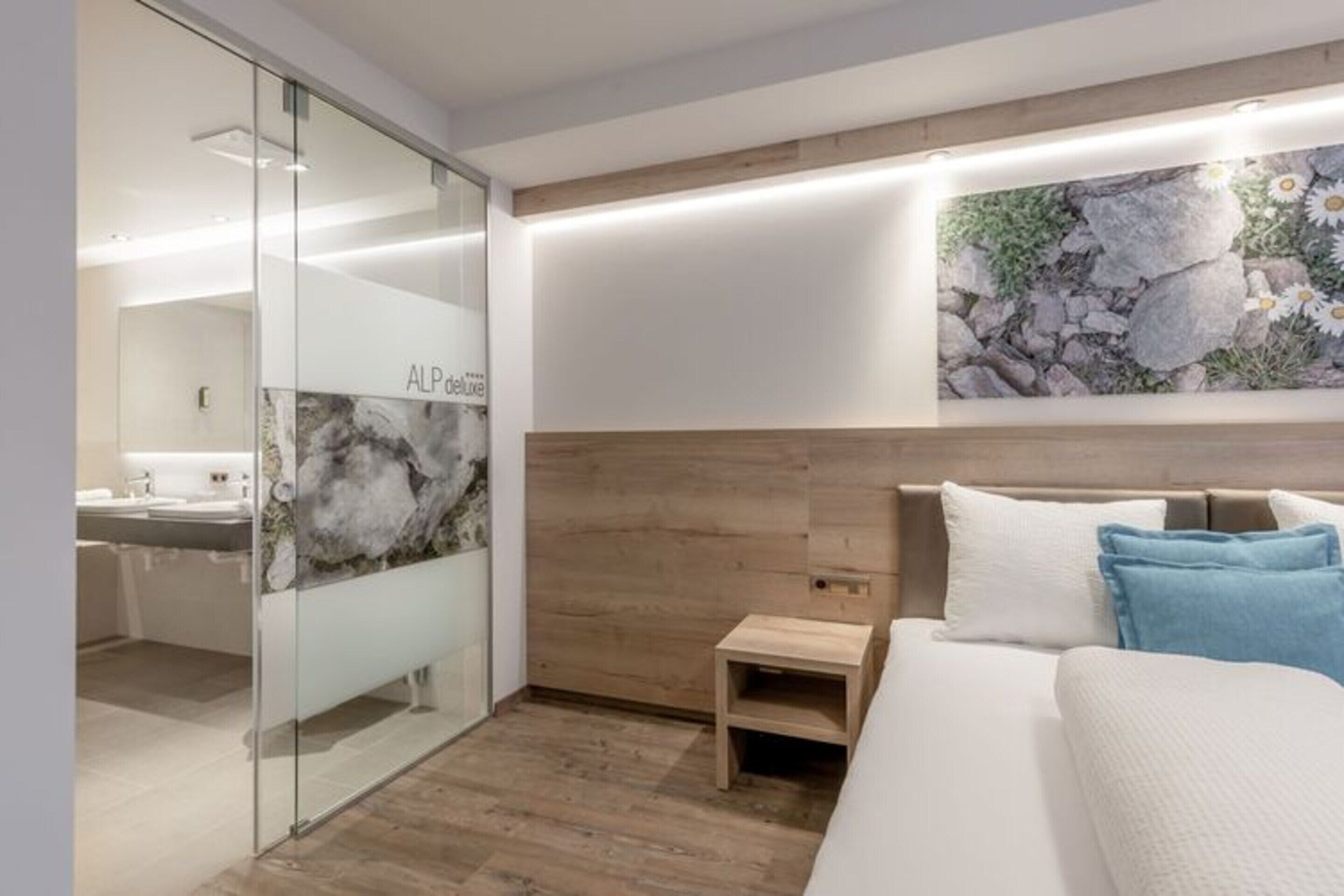 A modern bedroom with a bed, a small bedside table, a wall-mounted artwork, and a glass partition leading to a bathroom.