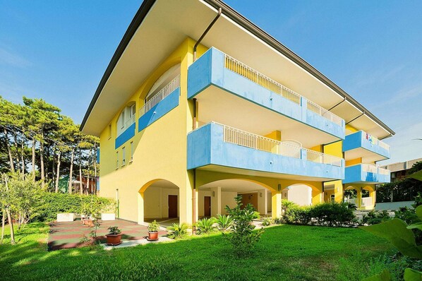 Un immeuble de deux étages avec une façade jaune et des balcons bleus, doté d’une entrée en arc de cercle et d’un petit espace jardin.