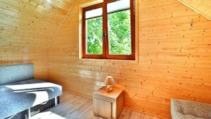 A wooden cabin room with a sloped ceiling, a window with a view of greenery, a small bedside table with a lamp, and a bench. - Comfortable Holiday Homes, Mielno (Mielno)