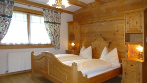 A wooden bedroom with a double bed, two bedside tables with lamps, a radiator, and a window with floral curtains. - Chalet Rabl, Westendorf (Westendorf)