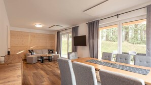 Ein moderner, gut beleuchteter Raum mit Holzboden, grau gepolsterten Möbeln, einem großen Tisch mit Stühlen, einem wandmontierten Flachbildfernseher und großen Fenstern mit Vorhängen.