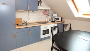 Ett kompakt kök med blå skåp, en vit ugn, en mikrovågsugn och en kaffebryggare. Det finns ett matbord med fyra stolar och ett fönster ovanför diskbänken.