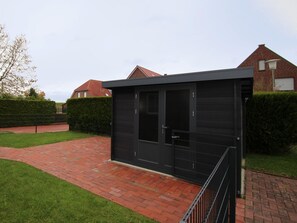 Un edificio moderno de color negro con techo plano, entrada protegida por una reja y un sendero de ladrillo que conduce a él.
