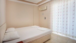 A single-bed room with a headboard, a bedside table, a wall-mounted air conditioner, and a window with patterned curtains. - Sevgi Tatil Sitesi (Alanya)