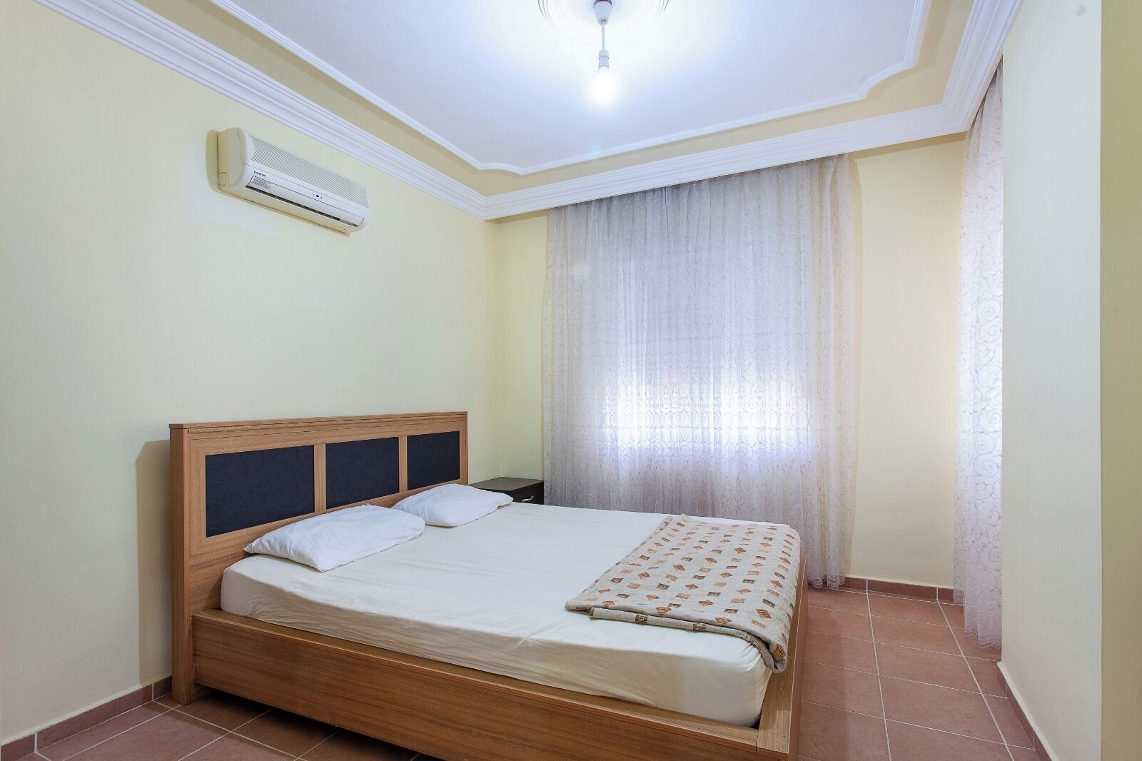 Un letto singolo con struttura e testiera in legno, un comodino, un condizionatore d'aria a parete e una finestra con tende leggere.