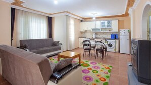 A compact living space with a sofa, a coffee table, a dining area with chairs, a kitchen with appliances, and a TV. - Sevgi Tatil Sitesi (Alanya)