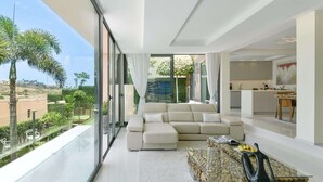 Ein modernes Wohnzimmer mit einer großen Couch, einem Glastisch und Blick auf einen Garten sowie auf die Stadt durch raumhohe Fenster.