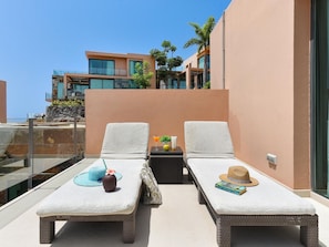 Two lounge chairs with towels, a small table with a drink, a hat, and a book on a balcony with a view of a modern building.