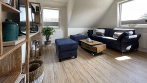 Ein modernes Wohnzimmer mit schrägverdeckter Decke, einem großen Fenster, einer blauen Sofagarnitur, einem Holztisch und einem Bücherregal mit verschiedenen Gegenständen.