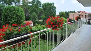 En balkong med ett metallräcke, ett kaklat golv och utsikt över en trädgård med röda blommor och gröna träd.