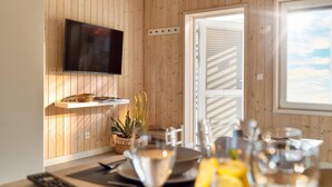 Ein Zimmer mit Holzwandverkleidung, an der Wand ist ein Fernseher angebracht, ein Esstisch mit Stühlen, einer Fernbedienung und einem Glas Wein.