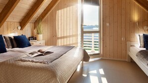 Ein gemütliches Schlafzimmer im Dachgeschoss mit schrägen Wänden, Holzboden und einem großen Fenster, ausgestattet mit einem Bett, einem Buch und Kissen.