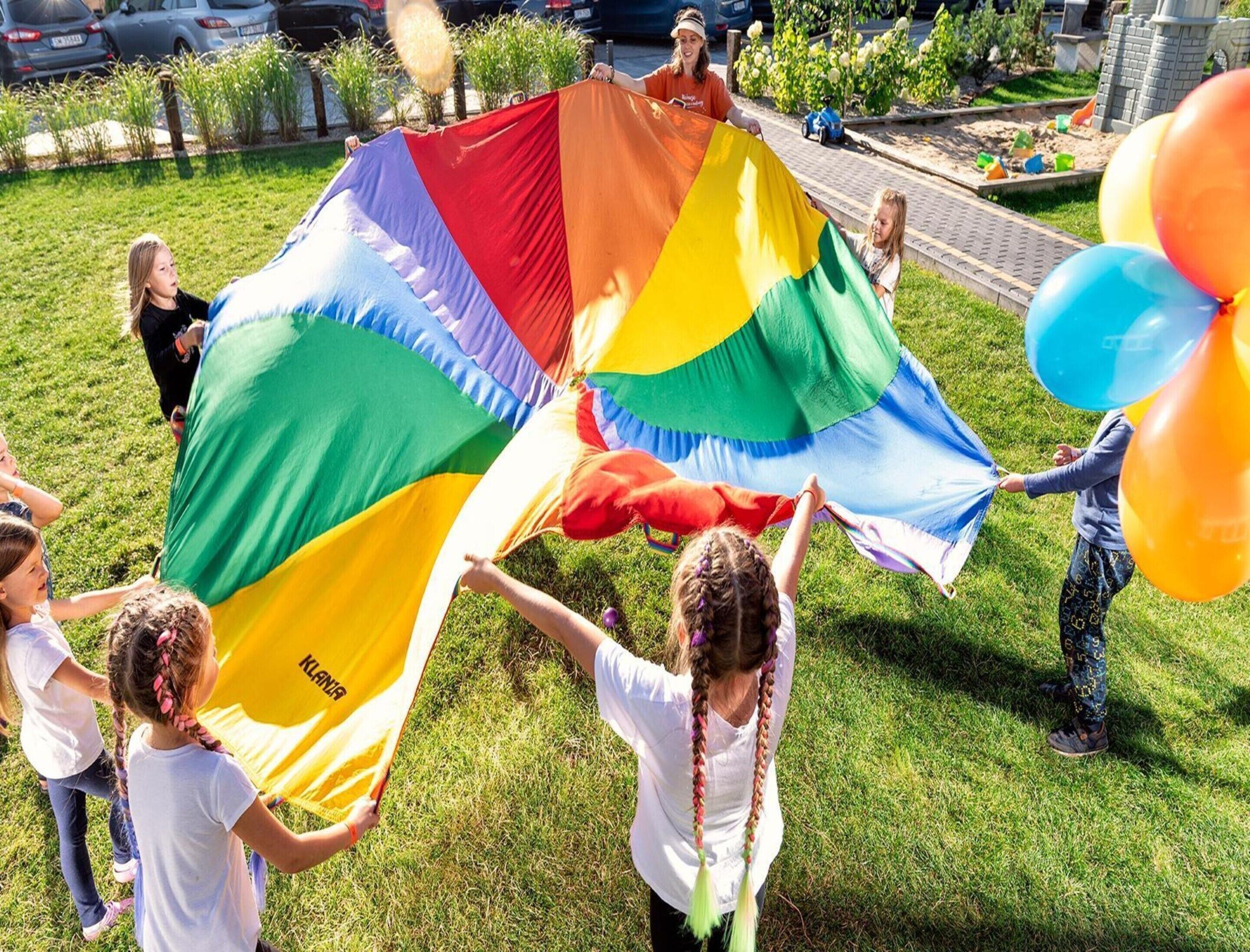 A group of children playing with a large rainbow-colored kite in a grassy outdoor area.