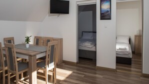 A compact room with a wooden dining table and chairs, a flat-screen TV mounted on the wall, a bed with a neatly made white duvet, and a framed picture of a sunset over the sea. - 3-room Apartment on the Ground Floor, Summer Pool (Postomino)
