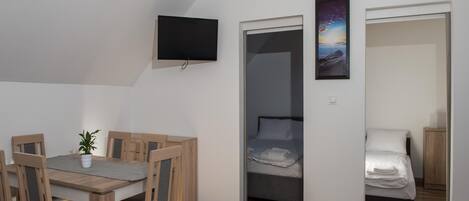 A compact room with a wooden dining table and chairs, a flat-screen TV mounted on the wall, a bed with a neatly made white duvet, and a framed picture of a sunset over the sea.