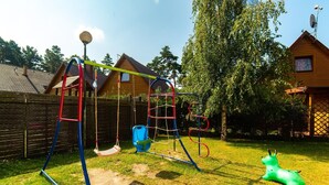 A playground with swings and a climbing structure in a backyard with a wooden fence and houses in the background. - 3-room Apartment on the Ground Floor, Summer Pool (Postomino)