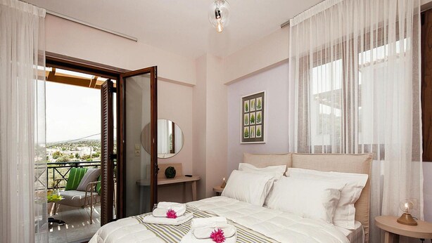 A hotel room with a large bed, a bedside table, a mirror, a window with sheer curtains, and a balcony with a chair and table. - Holiday Villa With hot tub in Adele (Rethymno)
