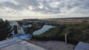 A coastal landscape with a stone wall, a wooden deck, and a distant view of a bridge and buildings. - Ferienwohnung in St. Peter-ording (St. Peter-Ording)