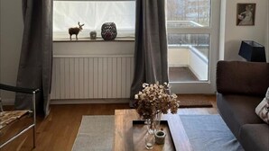 Un salon moderne doté d’une grande fenêtre, d’une table basse en bois sur laquelle se trouvent un vase et un plateau, d’un canapé marron et d’un radiateur placé sous la fenêtre.