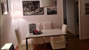 Un coin repas avec une table blanche, des chaises et un luminaire suspendu. Des tableaux encadrés ornent le mur, et une porte donne accès à une autre pièce.