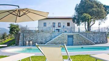 A pool area with lounge chairs, a stone wall, and a building with a staircase leading to a door.