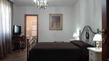 A hotel room with a bed, a television, a chandelier, a painting, and a desk with a lamp.