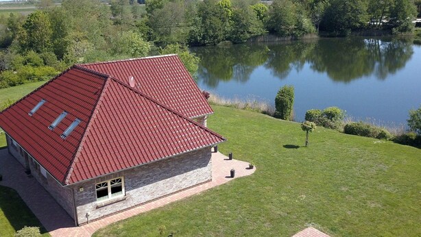 Ein Haus mit rotem Dach, steinernen Fundamenten und einer gepflasterten Auffahrt, die zu einer Rasenfläche mit Teich führt.