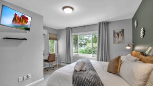 A modern bedroom with a large bed, a flat-screen TV mounted on the wall, a desk with a chair, and a window with curtains. - Gondola House | Hot tub, Arcade, Walkable Getaway, Fun Central! Game Room, Hot Tub, Steps to Village... (Ellicottville)