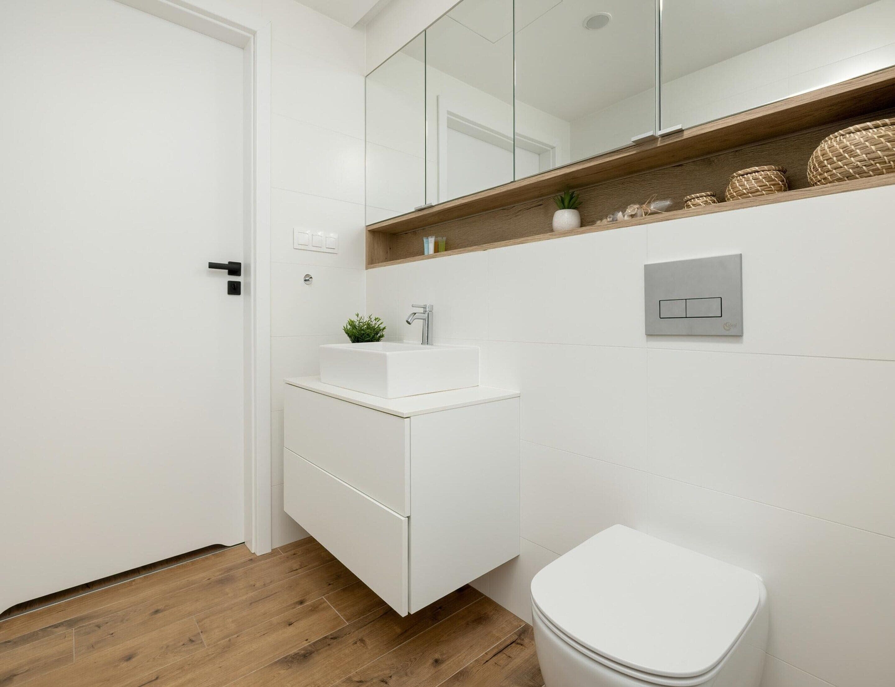 A modern bathroom with a white sink, a mirror above it, and a toilet. There is a wooden shelf with decorative items above the sink.