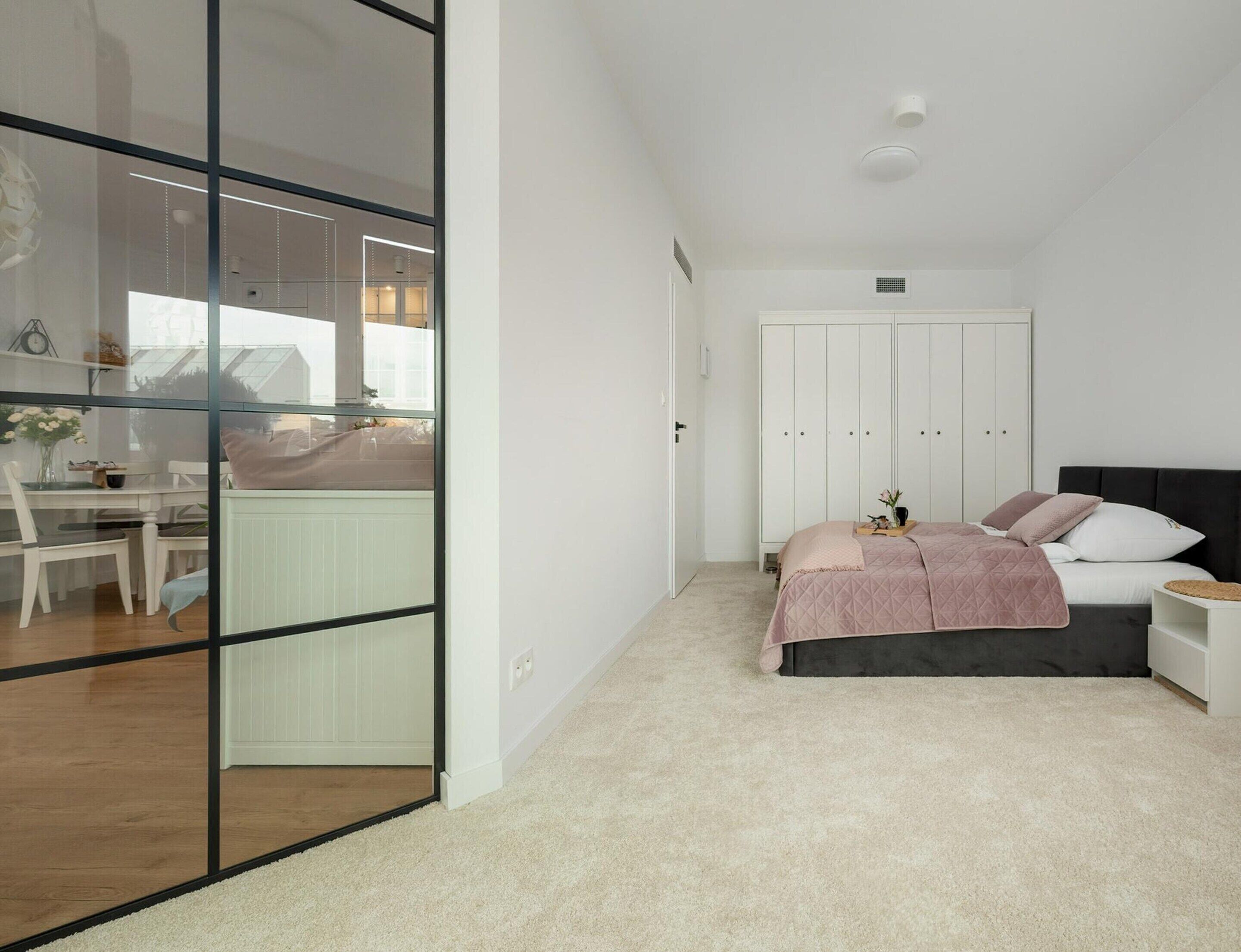 Ein Schlafzimmer mit Bett, Nachttisch und Kleiderschrank. Durch eine Glastrennwand ist eine Essecke mit Tisch und Stühlen zu sehen.