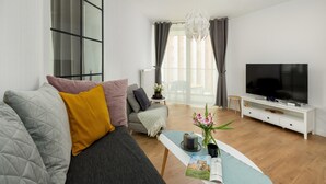 A modern living room with a sofa, a coffee table with a magazine, a TV, and a large window with curtains. - Apartment Close to the Sea, With Pool and Sauna (Rewal)