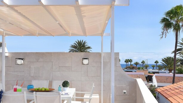 A covered outdoor seating area with a table set for two, a potted plant, and a view of the sea and palm trees. - Central Apartment With Spectacular Terrace-formerly TUI Ferienhaus (San Bartolomé de Tirajana)
