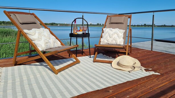 Two wooden lounge chairs with white cushions, a small table with a basket of fruit and glasses, and a straw hat on a wooden deck with a view of a lake. - Houseboat in Lazy by Lake Jamno and Sea (gmina Sianów)