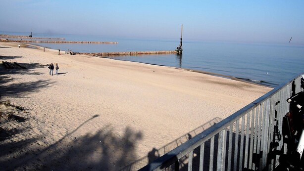 En strand med ett trästaket, stilla vatten och en brygga en bit bort.