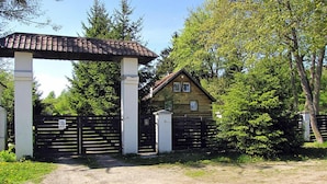 Une maison rustique en bois, avec un toit de tuiles, entourée d’une végétation luxuriante et dotée d’une entrée fermée par un portail.